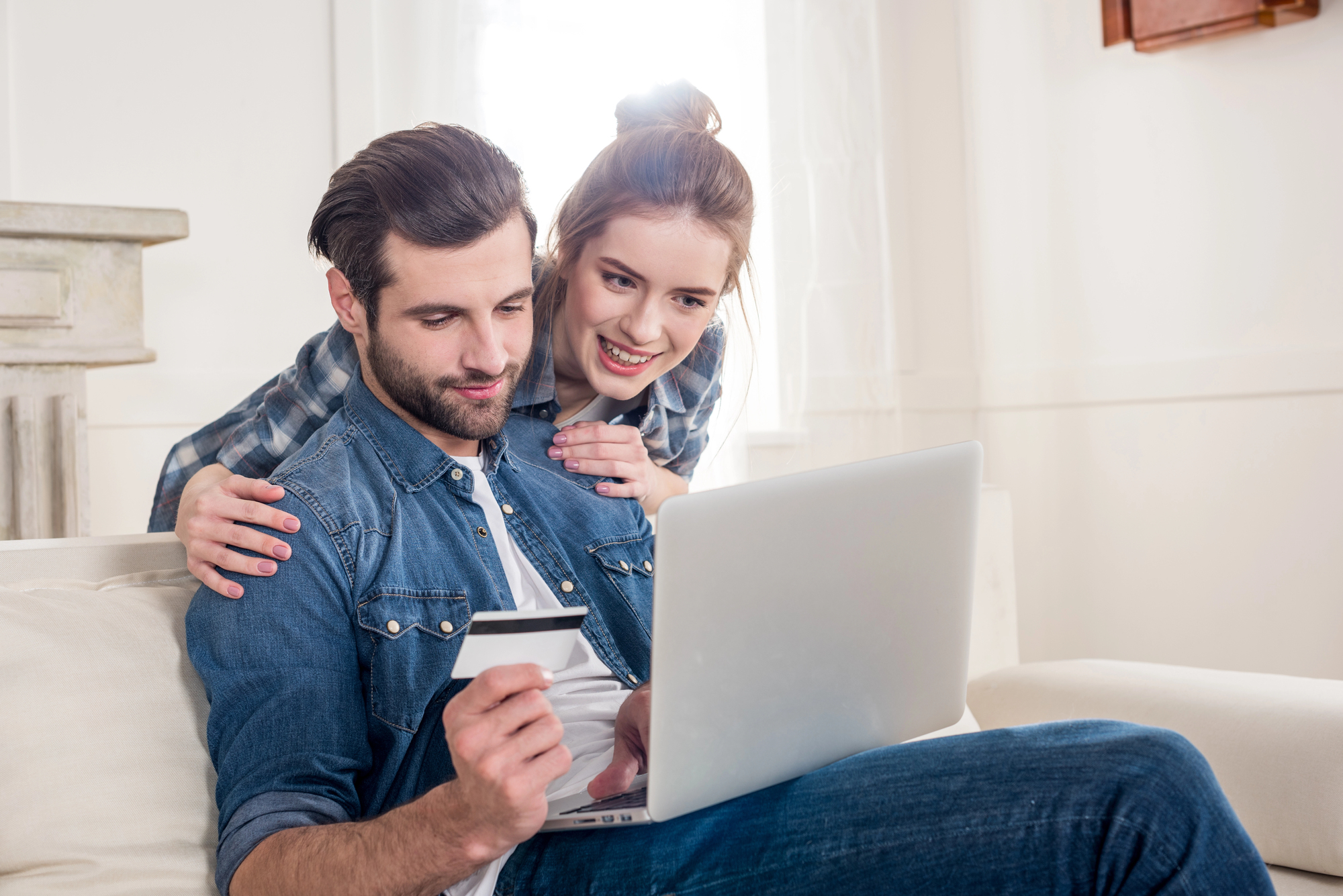 A man sitting on a couch uses a laptop while holding a credit card. A woman stands behind him with her arms around his shoulders, smiling and looking at the screen. Both are casually dressed in denim outfits. The room is bright and modern.