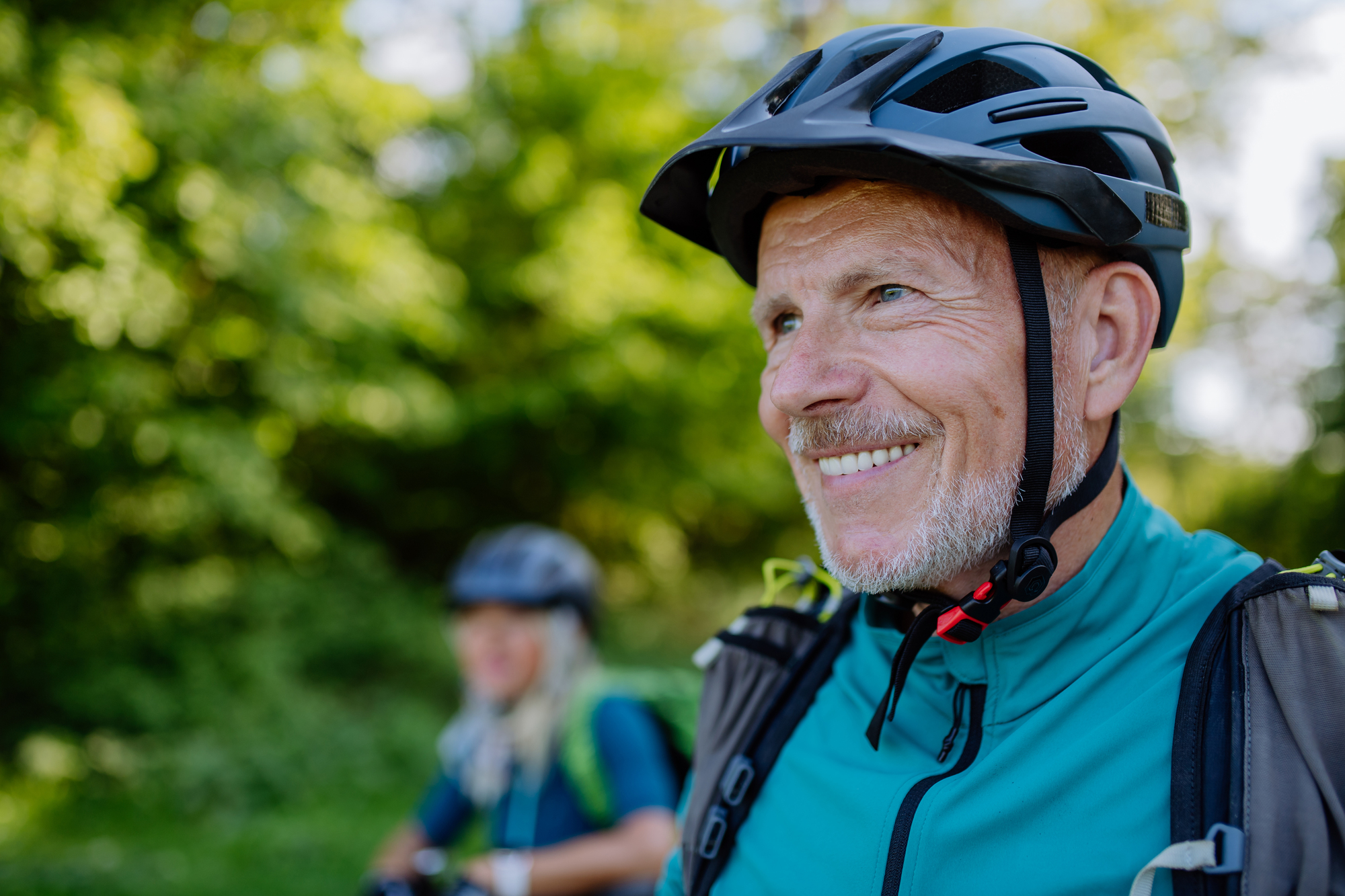 A smiling older man wearing a helmet and a green jacket is biking on a trail. A woman in the background is also biking, surrounded by lush green trees on a sunny day.