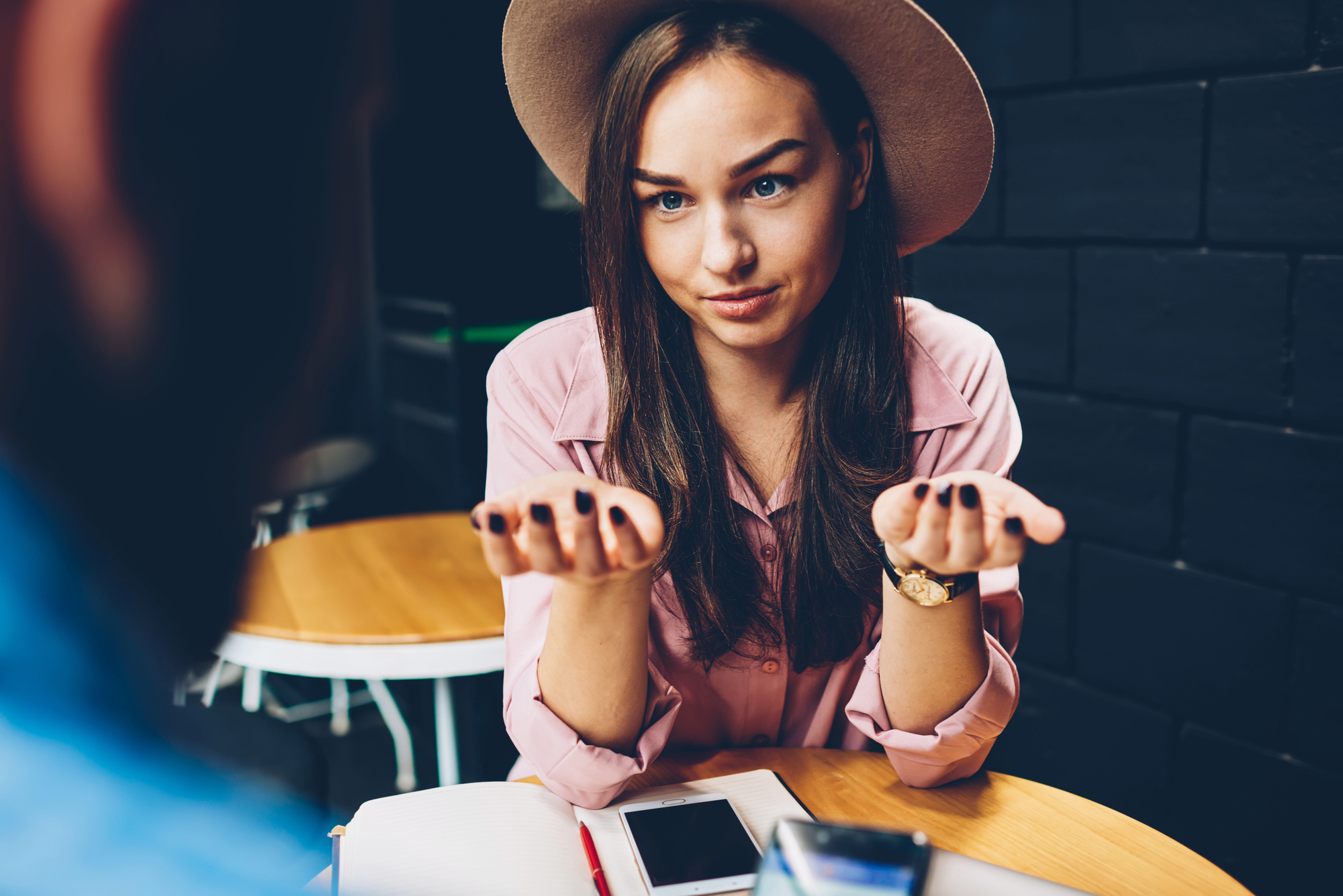 A woman in a hat and pink shirt sits at a table, gesturing with her hands as if in conversation. She looks engaged and expressive. A smartphone and notebook are on the table. The setting appears to be a casual cafe or meeting space.