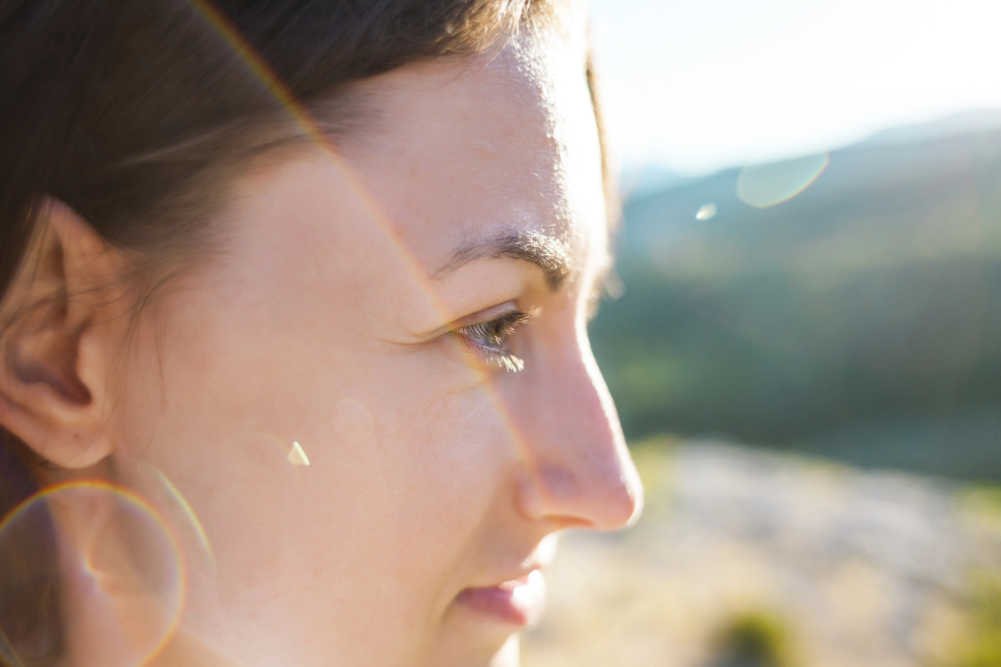 Close-up of a woman's face in profile, with sunlight creating lens flares and a gentle glow. The background appears to be a blurred natural landscape, suggesting an outdoor setting.