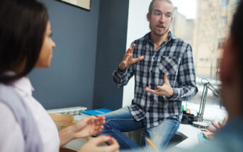 A bearded man in a plaid shirt is sitting on a desk, gesturing with his hands while talking. Two people are listening to him attentively. The scene is set in a well-lit office space with a window in the background.