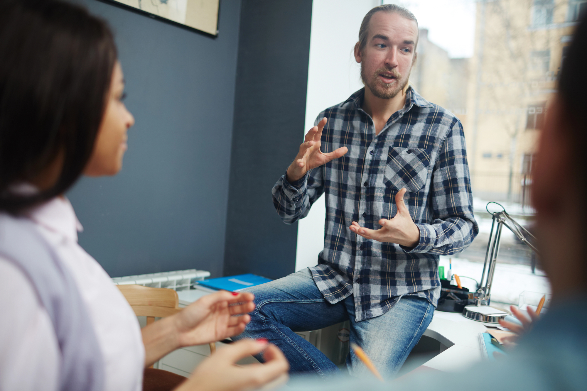 A bearded man in a plaid shirt is sitting on a desk, gesturing with his hands while talking. Two people are listening to him attentively. The scene is set in a well-lit office space with a window in the background.