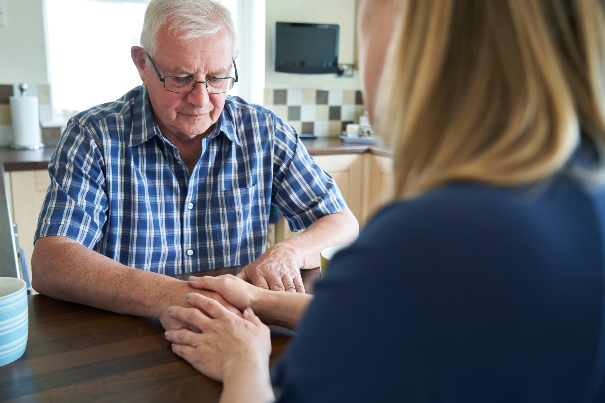 An elderly man in glasses and a plaid shirt sits at a table, holding hands with a woman facing him. They are in a kitchen, engaging in a comforting or serious conversation. A mug and household items can be seen in the background.