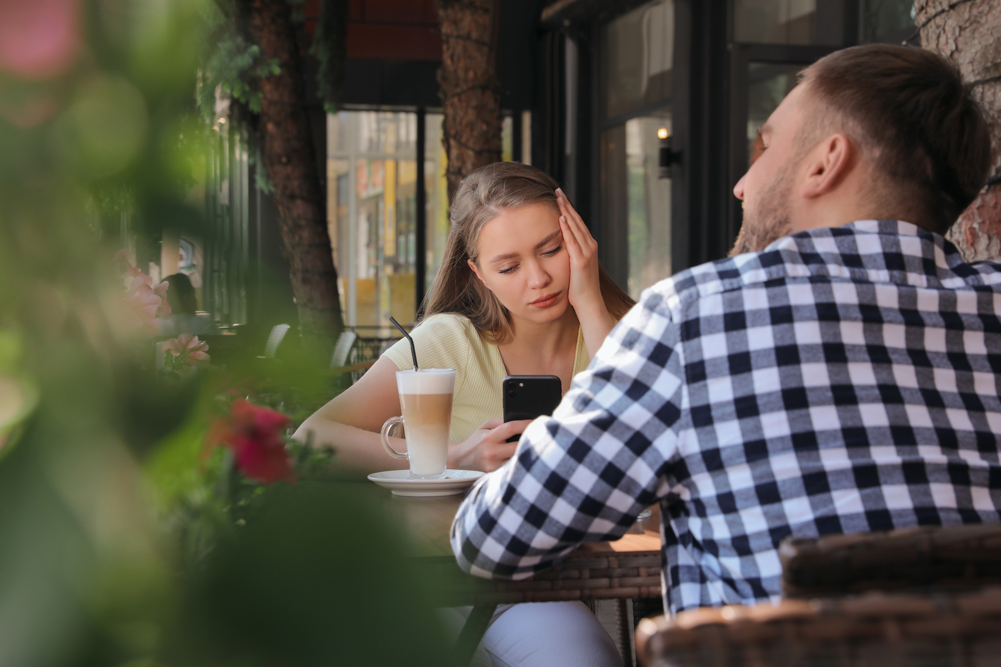 A woman sitting at an outdoor cafe table looks at her phone with a bored expression, while a man in a checkered shirt sits across from her, talking. A latte with a straw is on the table. Green foliage is visible in the foreground.