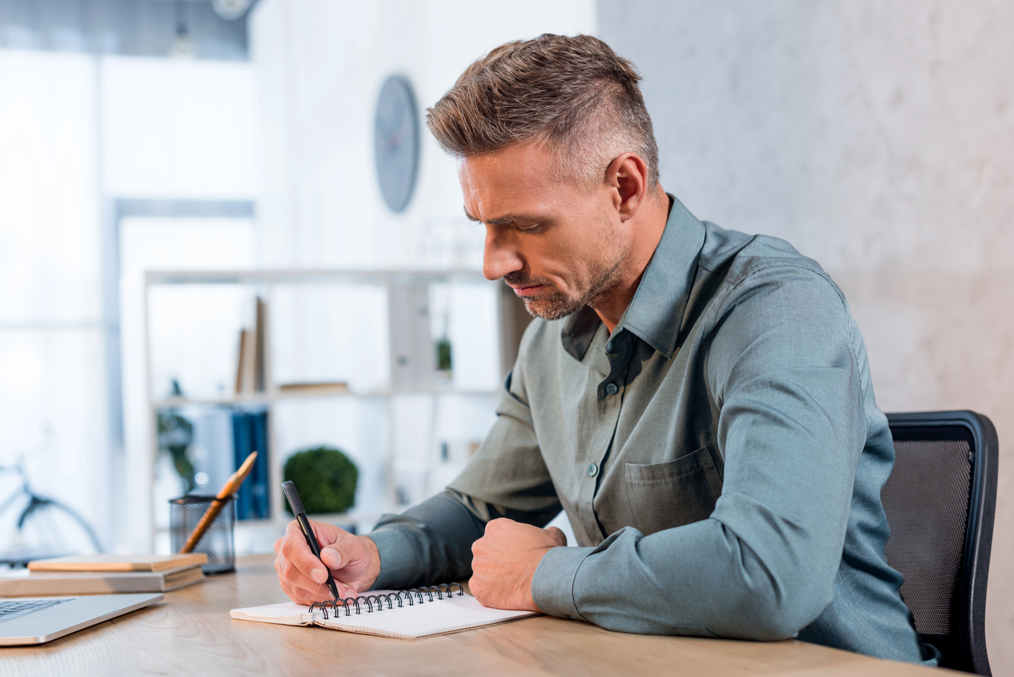 A man with short hair and wearing a gray shirt is sitting at a desk in an office, writing in a notebook. He appears focused, with a laptop and a potted plant in the background.