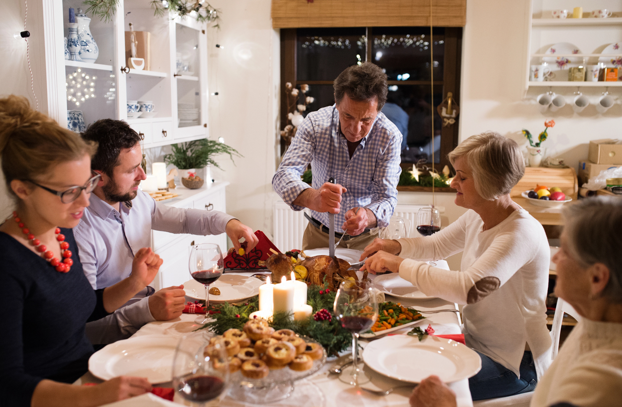 A family gathers around a dining table for a holiday meal. A man carves a roasted turkey while others enjoy wine and conversation. The table is decorated with candles, seasonal greenery, and various festive treats. The setting is cozy and warmly lit.