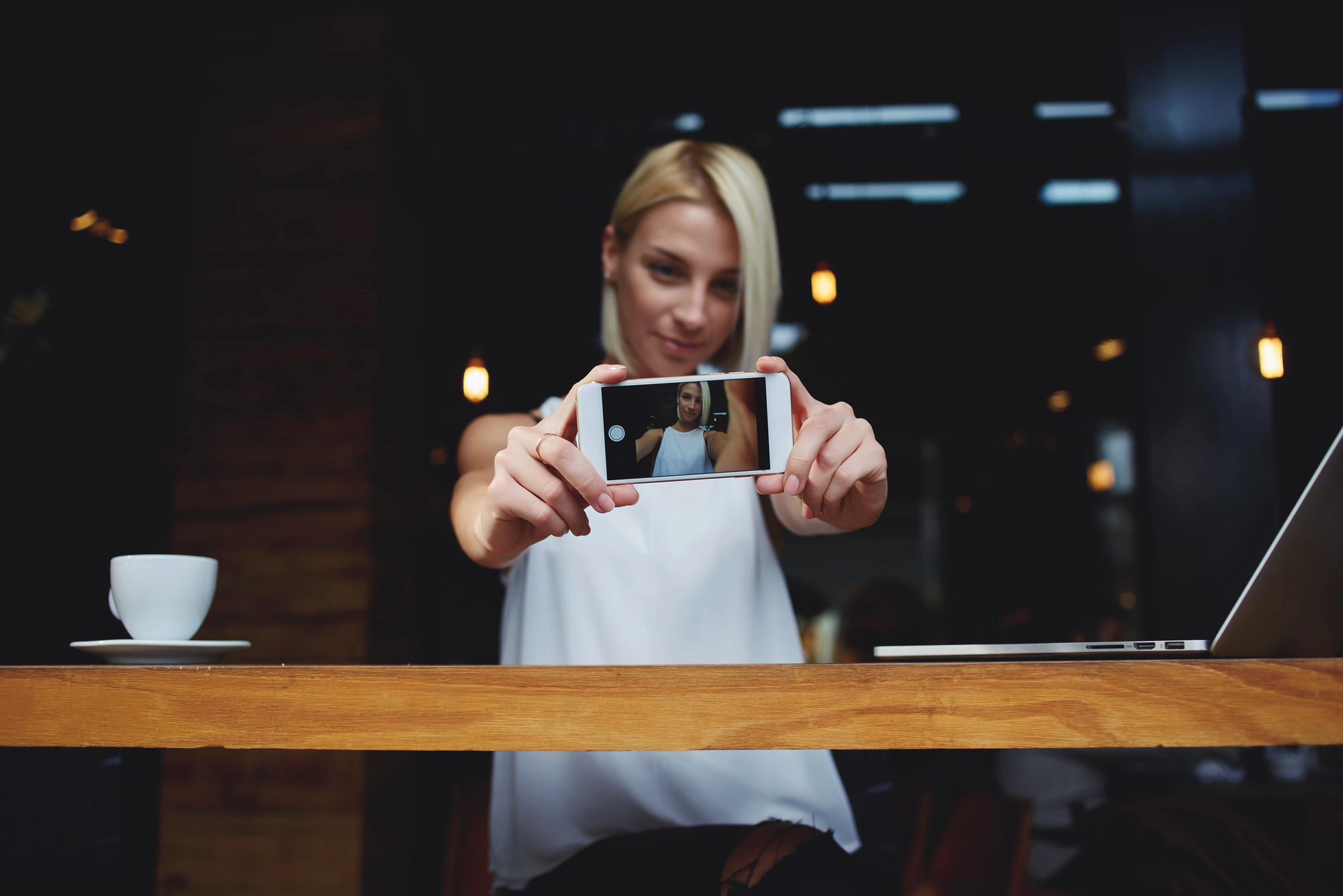 A person with blonde hair takes a selfie using a smartphone, holding it at arm's length. They are seated at a wooden table with a white coffee cup and an open laptop in a dimly lit room.