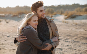 A couple stands on a sandy beach, wearing coats and smiling as they embrace. The background features dunes and softly blurred vegetation under a clear sky. The overall tone is warm and serene.