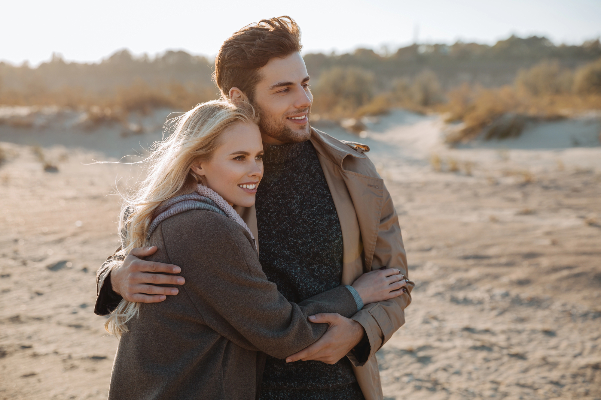 A couple stands on a sandy beach, wearing coats and smiling as they embrace. The background features dunes and softly blurred vegetation under a clear sky. The overall tone is warm and serene.