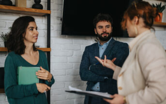 Three people are having a discussion in a modern office setting. A woman in a green top holds a book, a man listens with arms crossed, and another woman gestures while speaking. Shelves with decor and a TV are in the background.