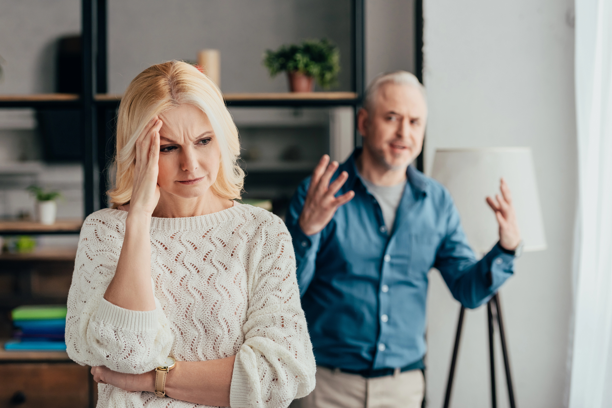 A woman with a hand on her forehead looks concerned while standing in a room. In the background, a man gestures with raised hands, appearing frustrated. They are in a domestic setting with shelves and a lamp visible.