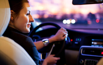 A woman driving a car at dusk, smiling and looking out the window. The dashboard is illuminated, and blurred lights are visible through the windshield, suggesting an urban setting.