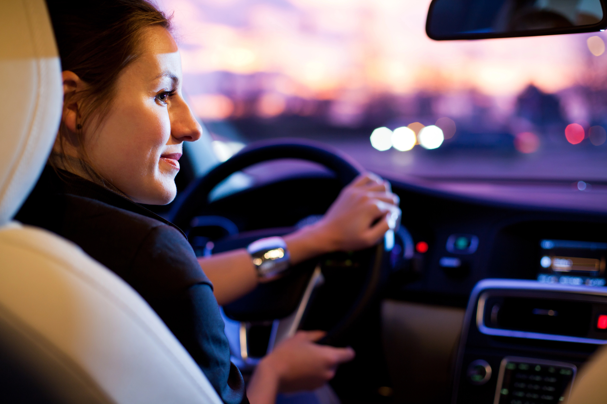 A woman driving a car at dusk, smiling and looking out the window. The dashboard is illuminated, and blurred lights are visible through the windshield, suggesting an urban setting.