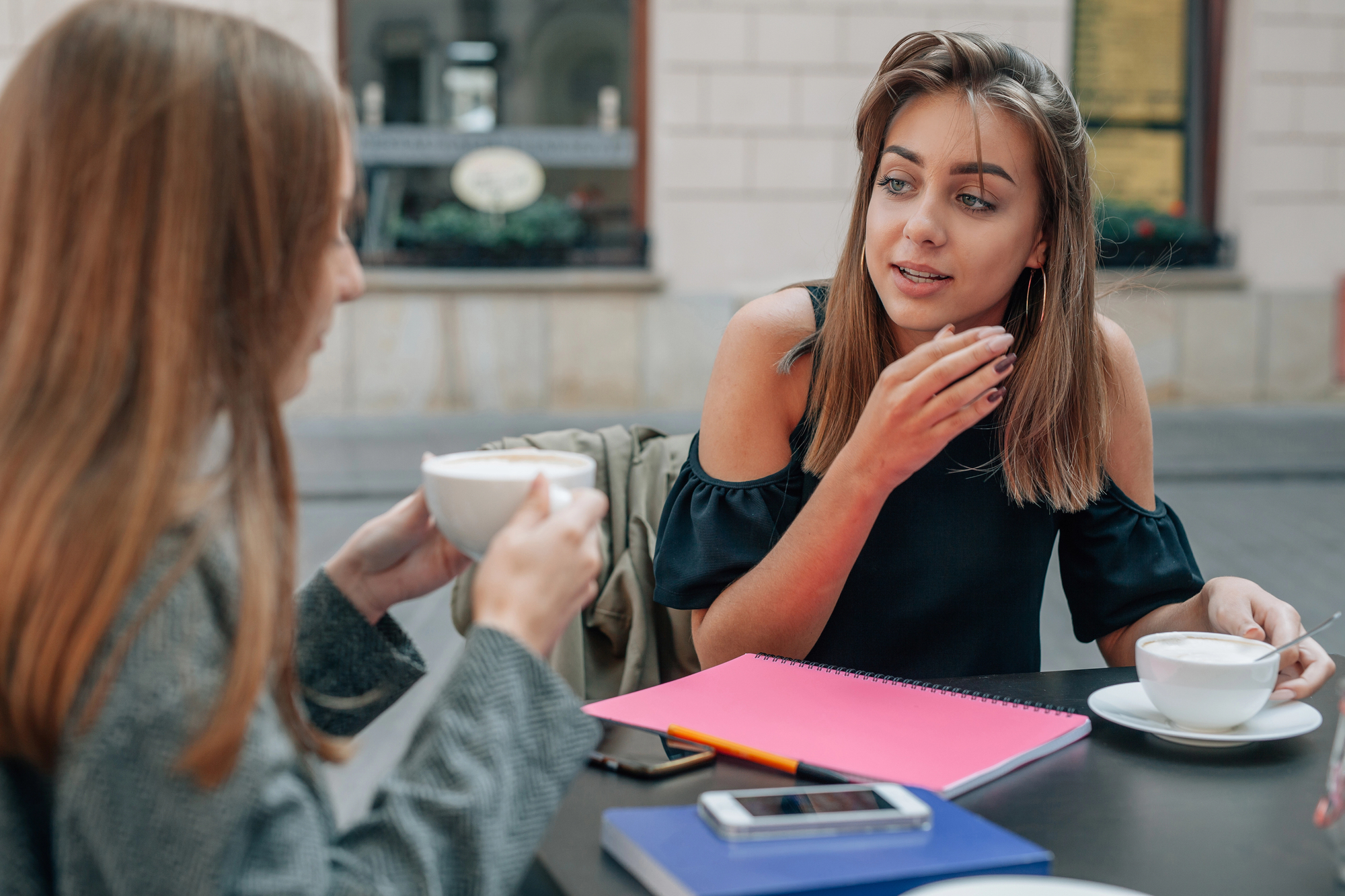 Two women sit at an outdoor café table, engaged in conversation. One gestures while holding a coffee cup, and the other listens with a cup in her hand. A pink notebook and a smartphone with earbuds are on the table.