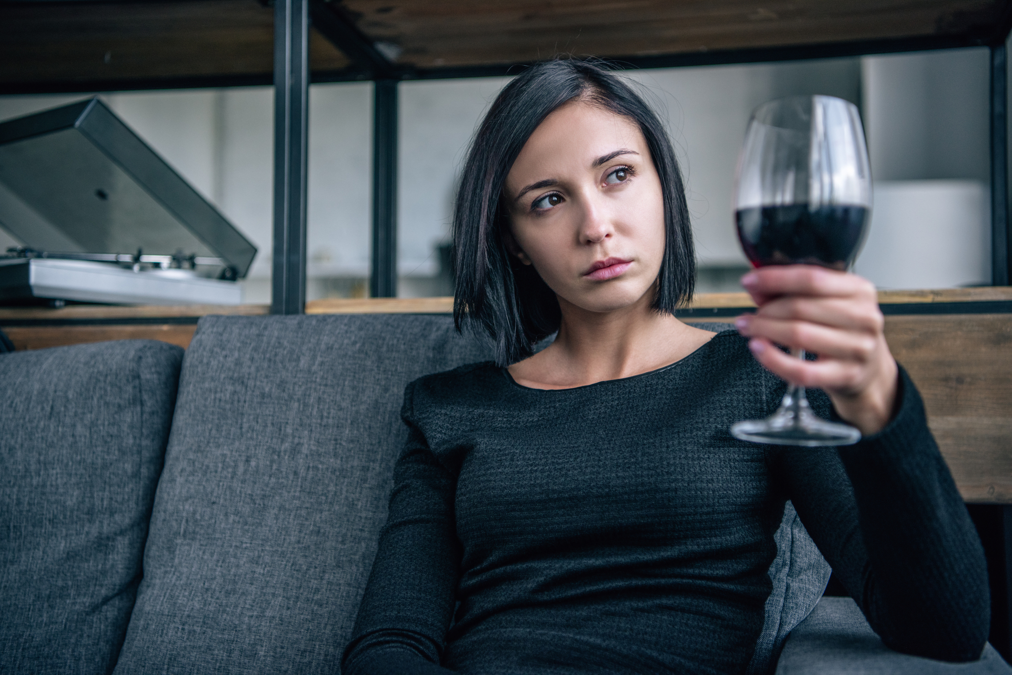 A woman with short dark hair sits on a gray couch, wearing a black long-sleeve top. She is holding a glass of red wine, gazing at it thoughtfully. A turntable is visible in the background on a shelf.