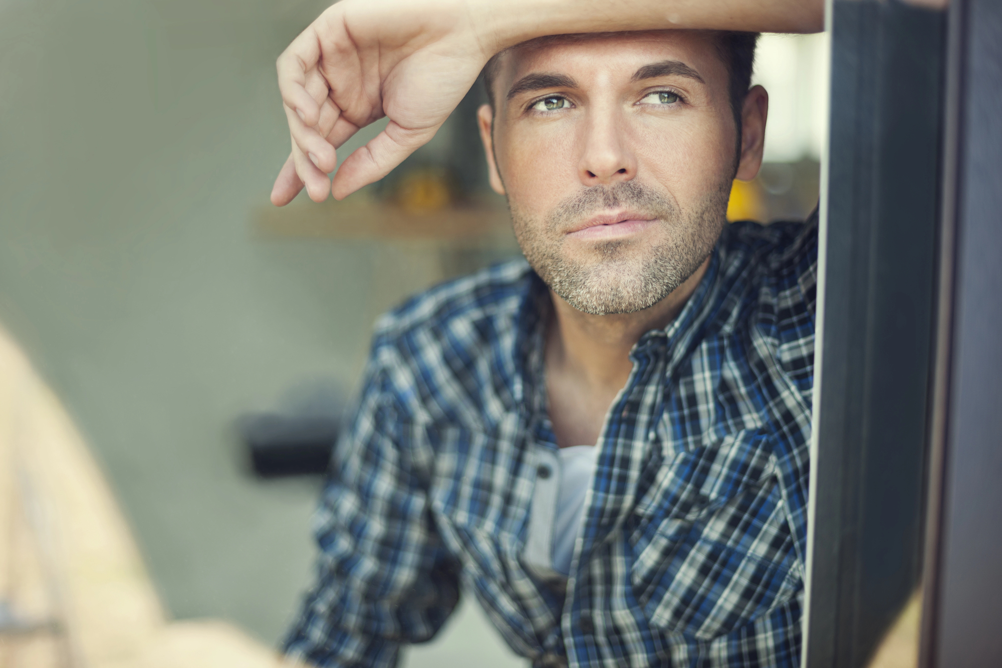 A man in a blue plaid shirt gazes thoughtfully out of a window. His right arm is raised, with his hand resting on his forehead. The background is softly blurred, suggesting an indoor setting.