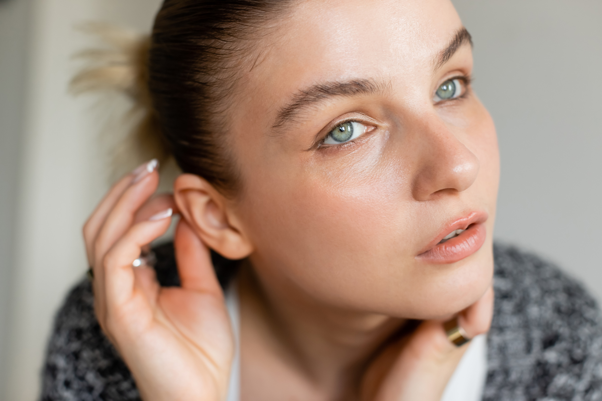 A woman with light-colored eyes and natural makeup looks towards the camera. She gently touches her ear with one hand, while the other is near her chin. Her hair is tied back, and she wears a gray sweater. The background is neutral.