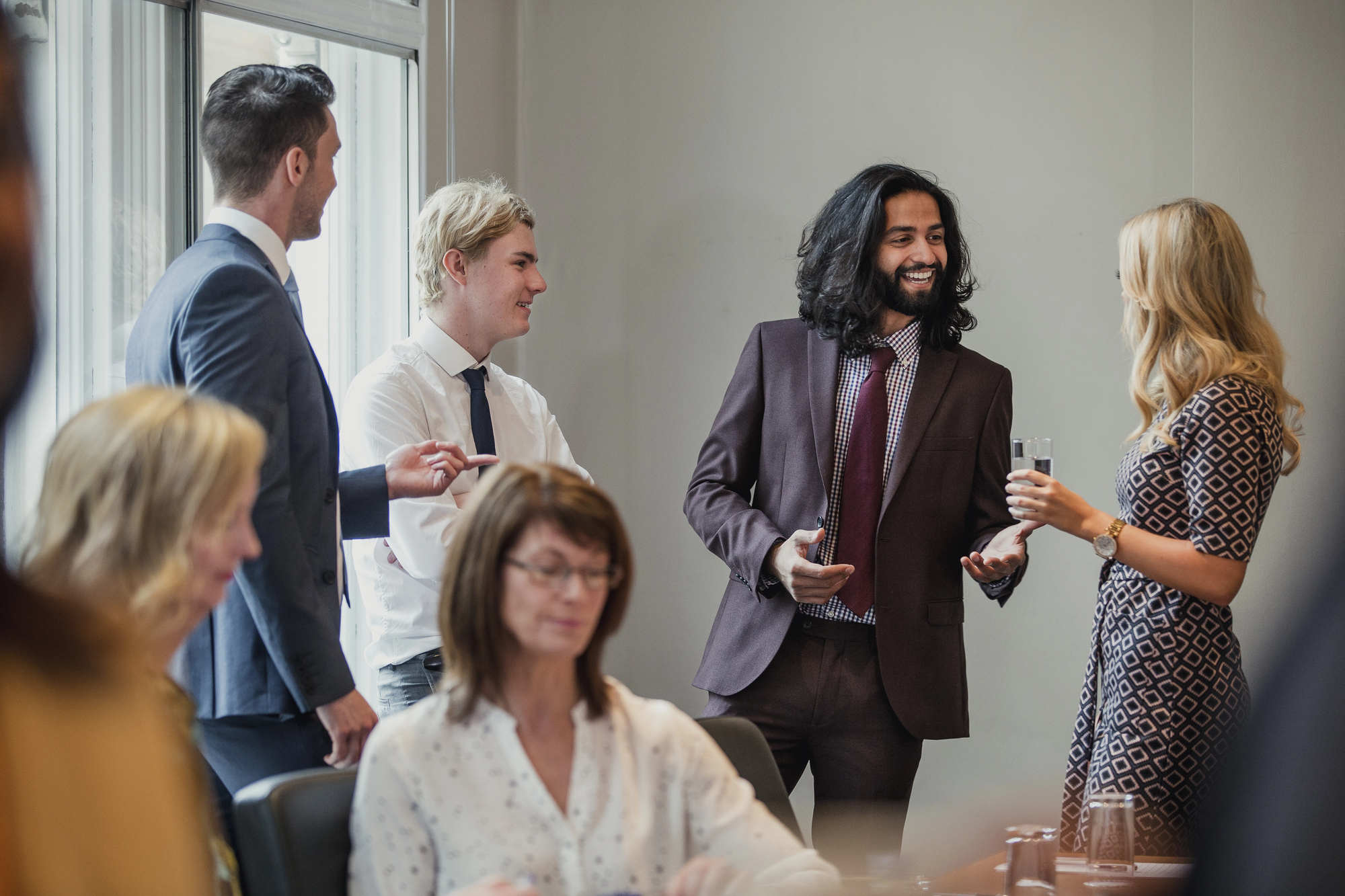 A group of people in business attire are engaging in conversation in an office setting. One person is holding a drink. Others are sitting and listening. The mood appears casual and friendly.