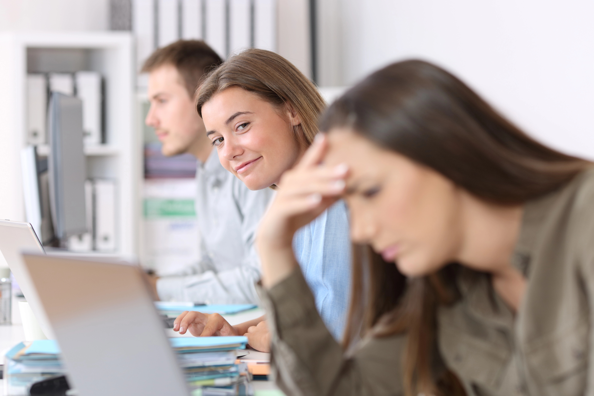 A woman smiles playfully at the camera while sitting at a desk next to two other colleagues working on laptops. The person in the foreground appears focused with their hand on their forehead. The background features office furnishings.