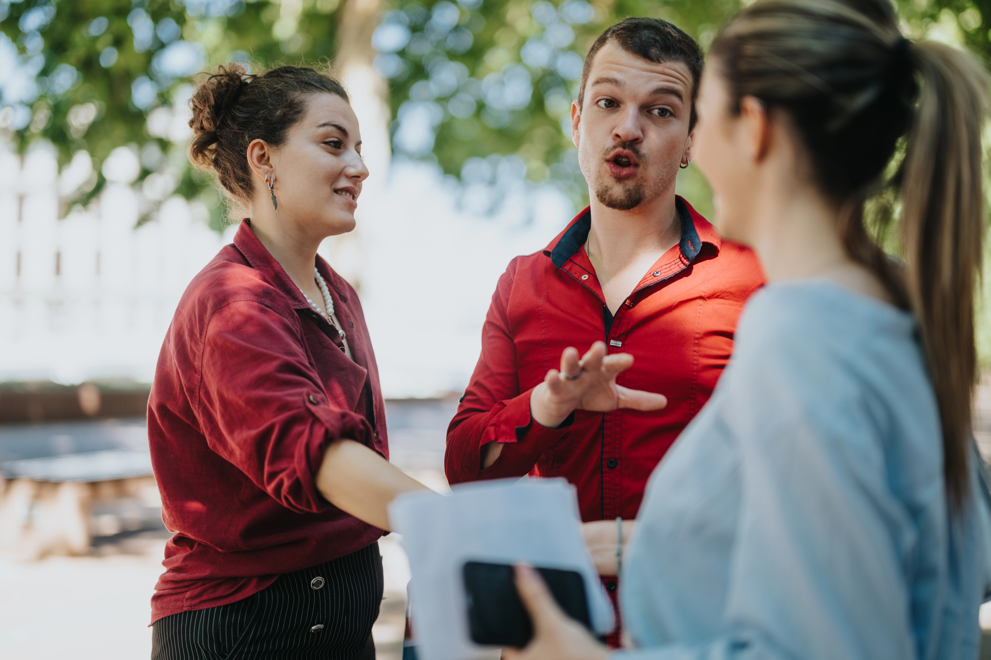 Three people are having a discussion outdoors. One man in a red shirt appears animated, gesturing with his hands. Two women, one holding papers and a phone, listen. They stand near trees, with sunlight filtering through the leaves.