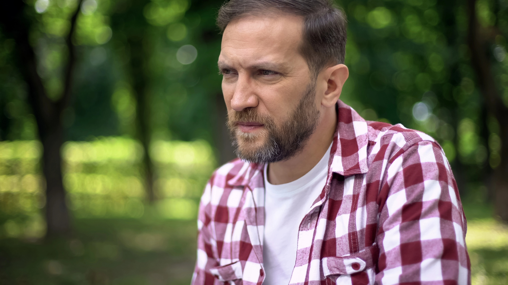 A man with a beard and short hair, wearing a red and white checkered shirt over a white t-shirt, is sitting outdoors in a park. The background is lush and green with blurred trees and foliage. He appears thoughtful.