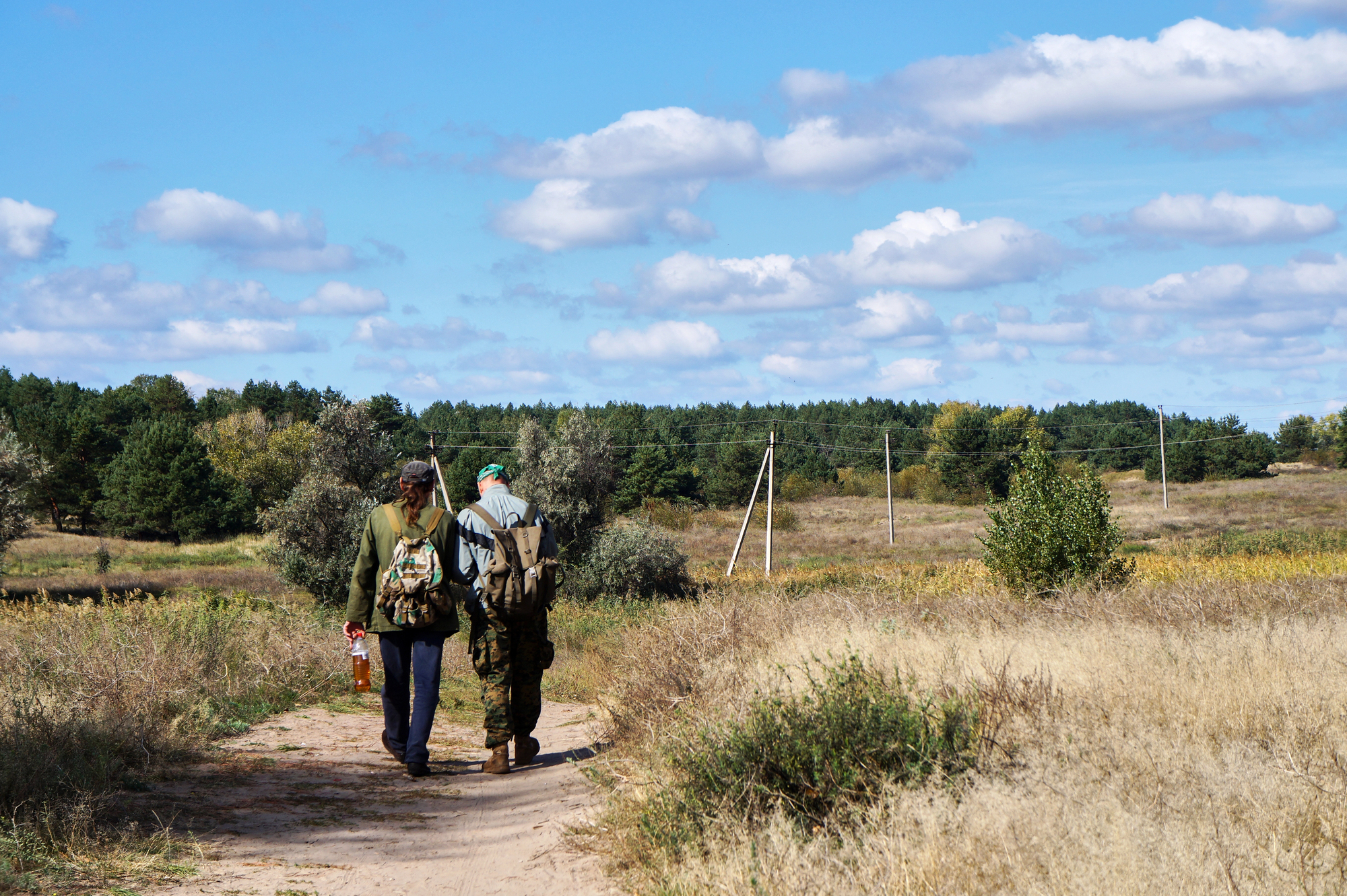 Two people wearing backpacks and outdoor clothing walk along a dirt path through a grassy, rural landscape. The sky is blue with scattered clouds, and trees line the distant horizon. One person holds a bottle in their hand.