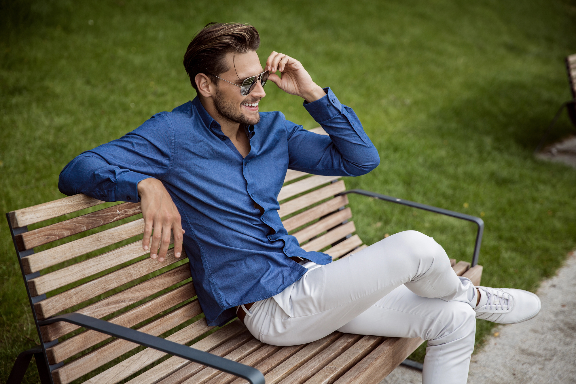 A man in a blue button-up shirt and white pants sits casually on a wooden park bench. He is wearing sunglasses, has his left arm resting on the back of the bench, and is smiling. The background features lush green grass.