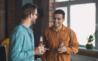 Two men are standing in a well-lit room, smiling at each other while holding beer bottles. One wears a blue shirt and sunglasses, the other an orange shirt. A large window and a potted plant are in the background.