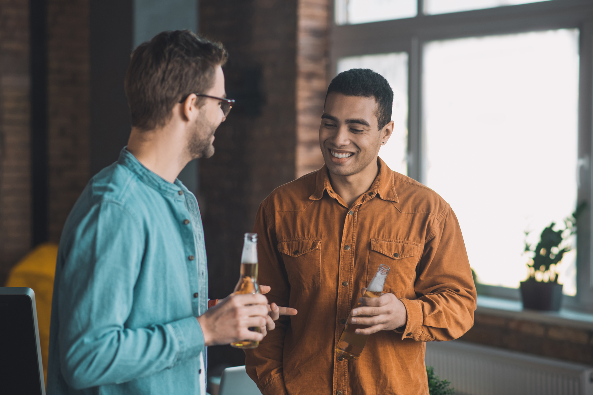 Two men are standing in a well-lit room, smiling at each other while holding beer bottles. One wears a blue shirt and sunglasses, the other an orange shirt. A large window and a potted plant are in the background.