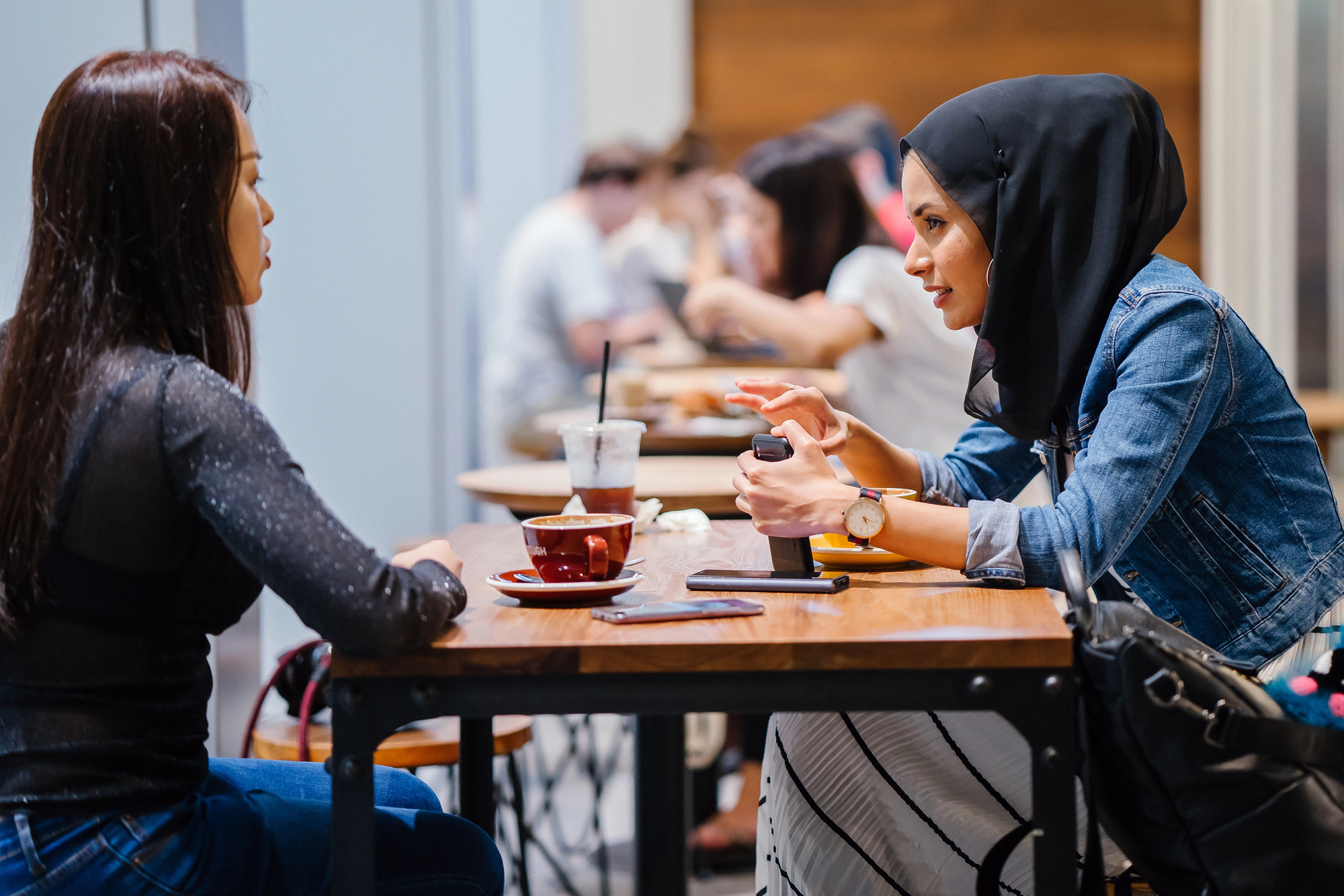 Two women sitting at a cafe table, engaged in conversation. One woman wears a hijab and denim jacket, while the other has long hair and a dark top. They have drinks in front of them, and there are blurred people in the background.