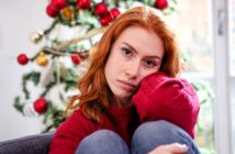 A red-haired woman in a red sweater sits pensively with her knees up, in front of a decorated Christmas tree adorned with red and gold ornaments. Bright natural light filters through a nearby window.