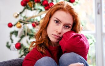 A red-haired woman in a red sweater sits pensively with her knees up, in front of a decorated Christmas tree adorned with red and gold ornaments. Bright natural light filters through a nearby window.