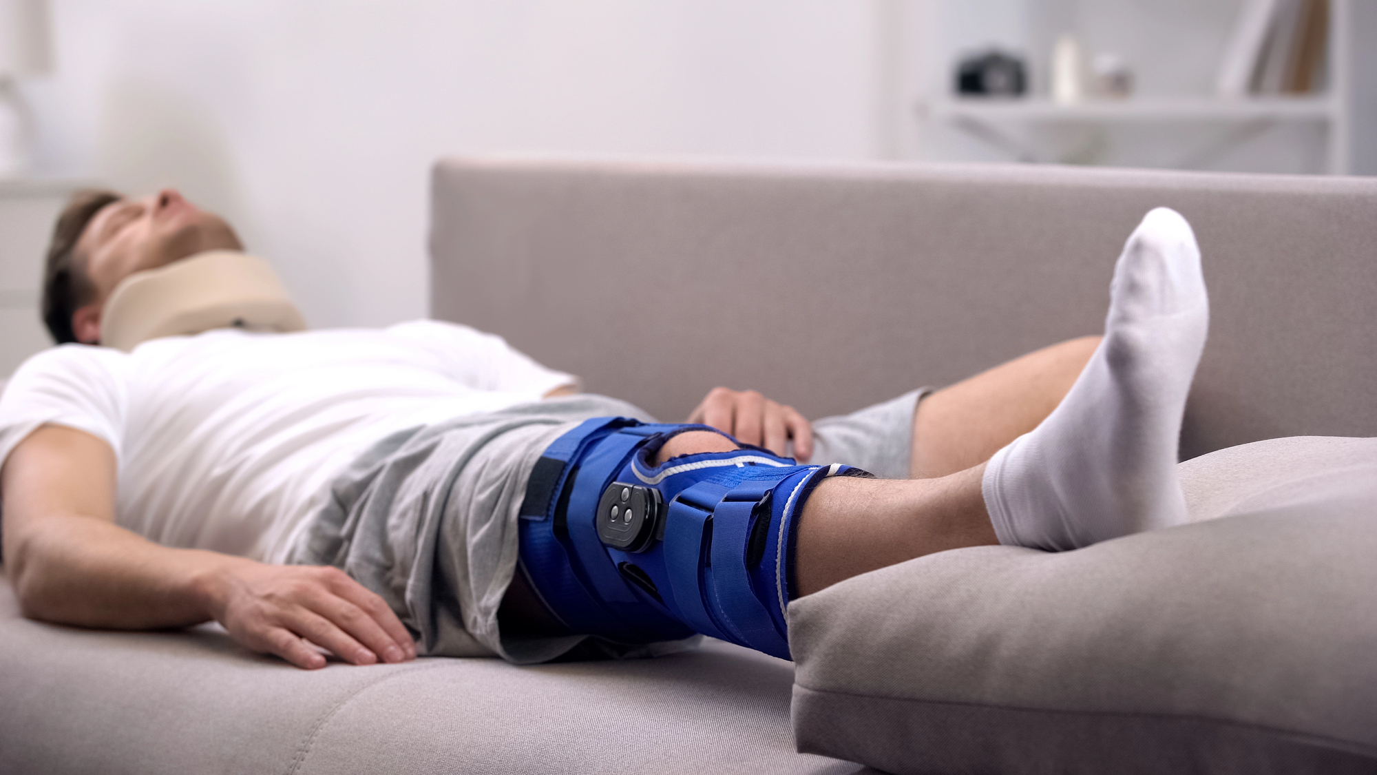 A person lying on a couch wearing a neck brace and a blue knee brace, dressed in a white t-shirt and gray shorts. Their legs are elevated on the arm of the couch, and they appear to be resting or asleep.