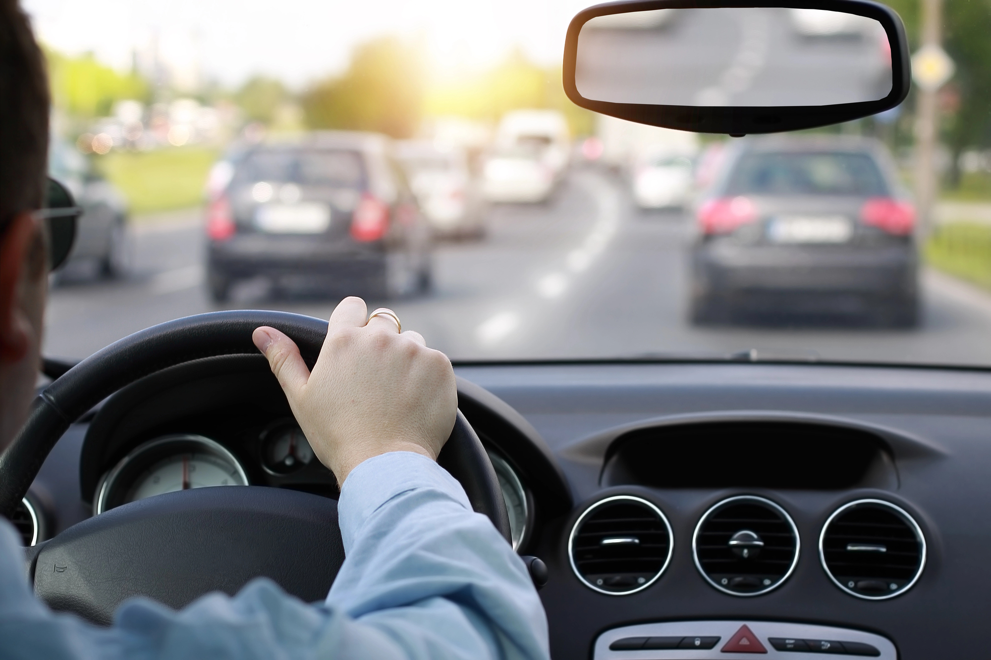 A person driving a car, holding the steering wheel with one hand. The view from inside the car shows traffic on a busy road with multiple vehicles ahead in the background, under a bright sky.