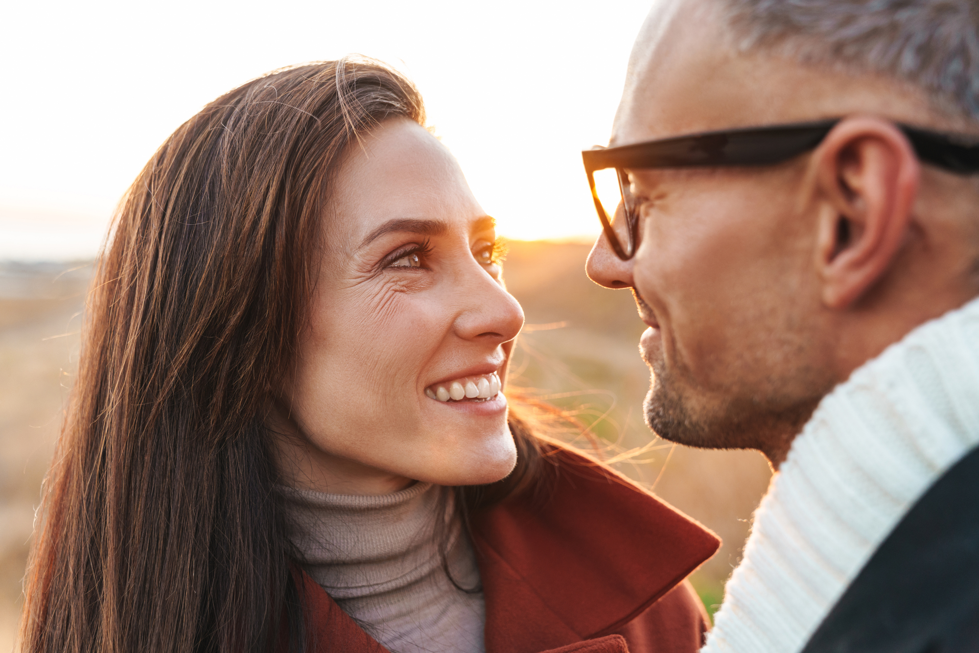 A couple facing each other smiles warmly against a softly lit outdoor backdrop at sunset. The woman has long brown hair and wears a red coat, while the man has glasses and wears a white scarf. Their expressions convey warmth and affection.