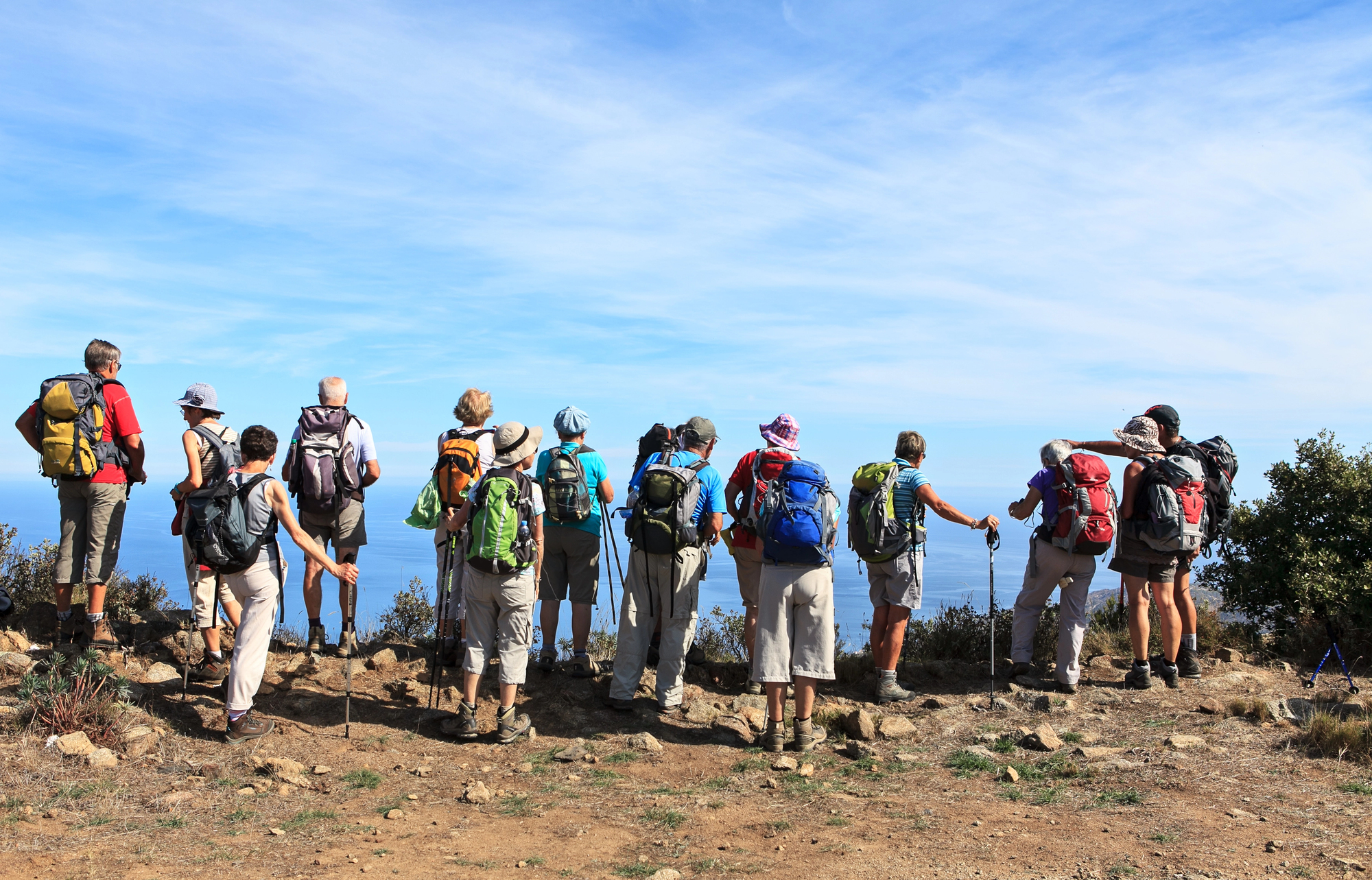 A group of hikers with backpacks stand on rocky terrain, facing a scenic view of the ocean under a clear blue sky. The hikers are dressed in light clothing and use hiking poles. Vegetation surrounds the area.