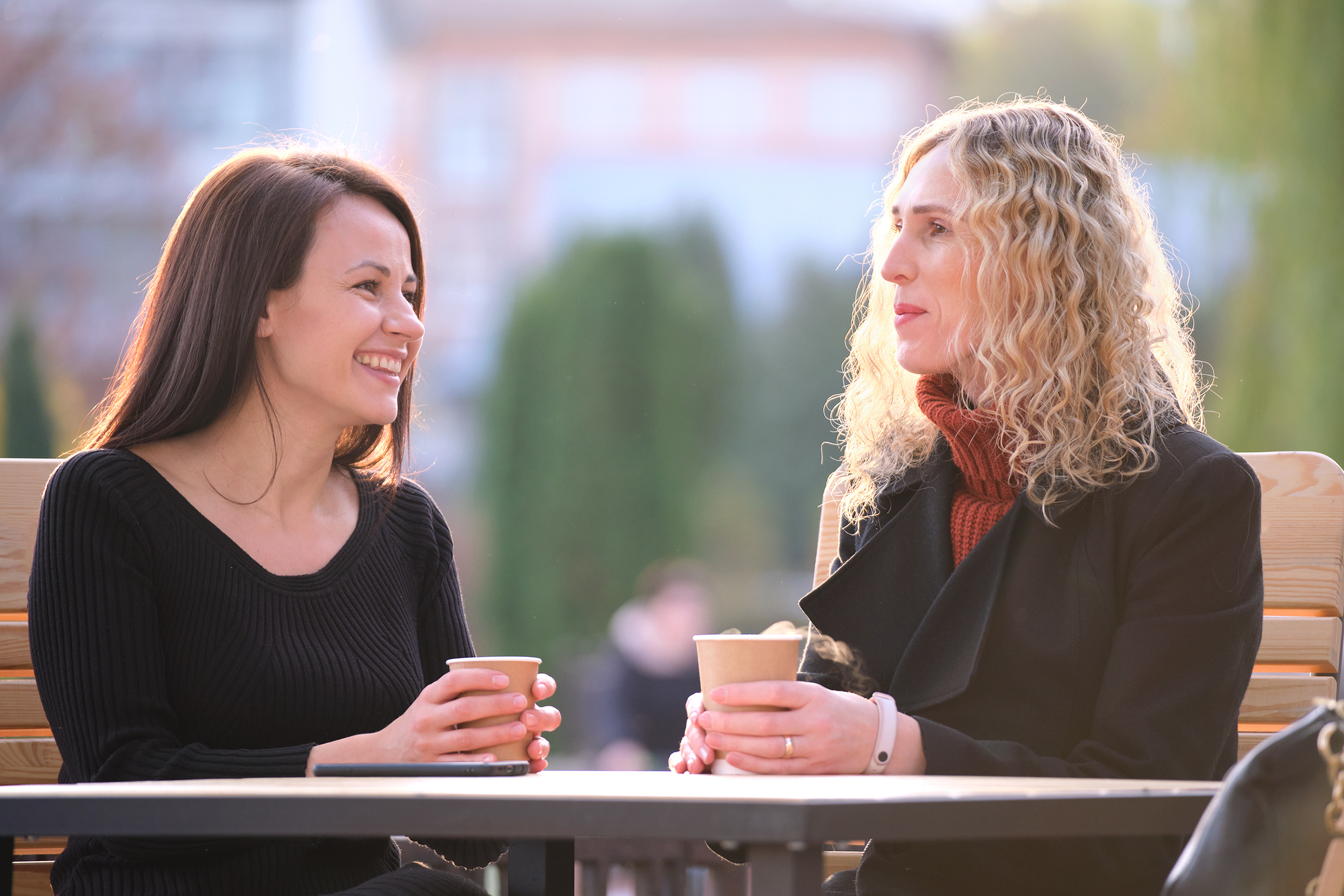 Two women sit at an outdoor table, smiling and holding coffee cups. One has long, dark hair and is wearing a black top, while the other has curly blonde hair and is wearing a black coat and maroon scarf. The background is blurred with greenery.