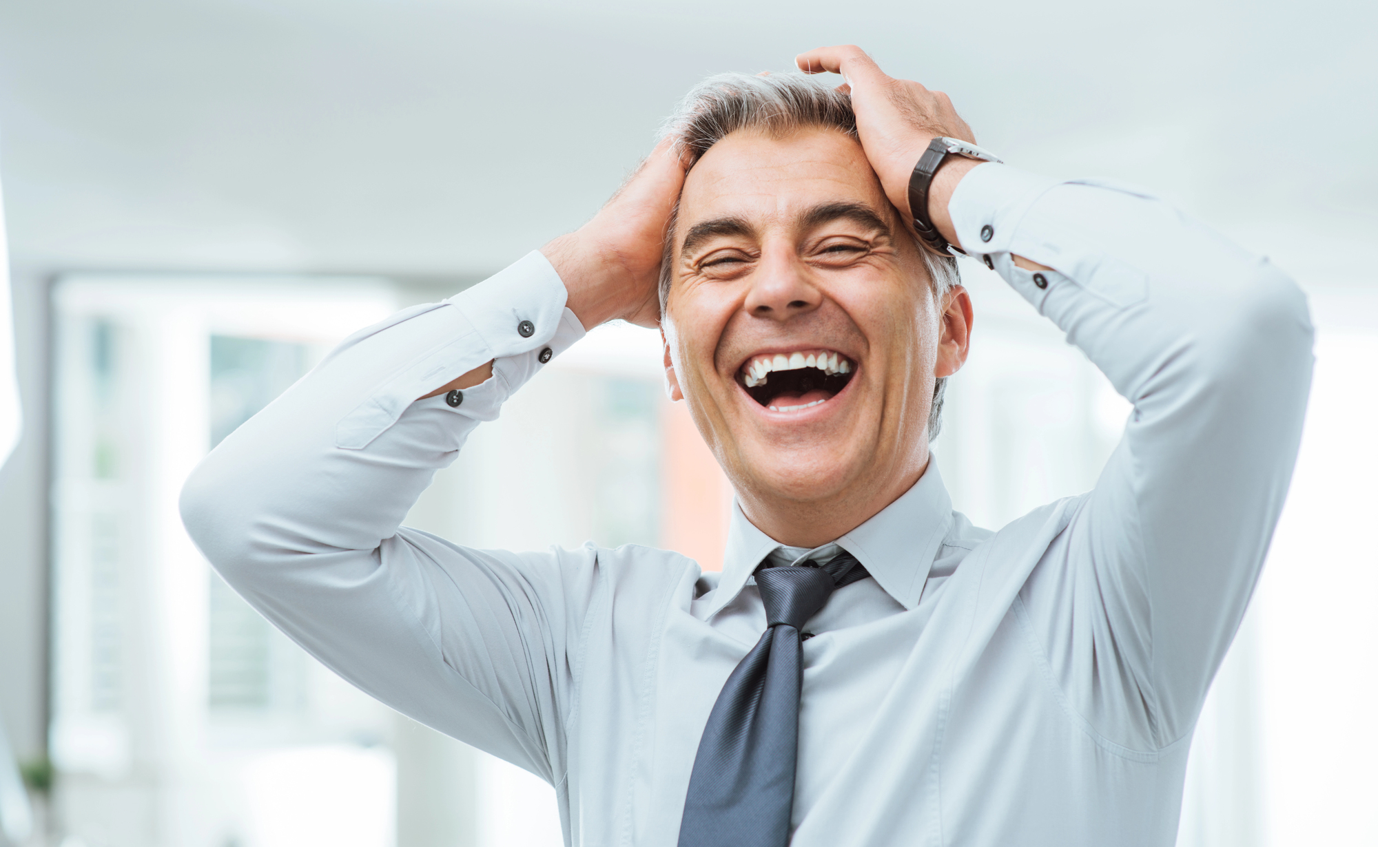 A man in a white shirt and tie is laughing heartily with his hands on his head. He appears to be standing indoors, and the background is softly blurred.