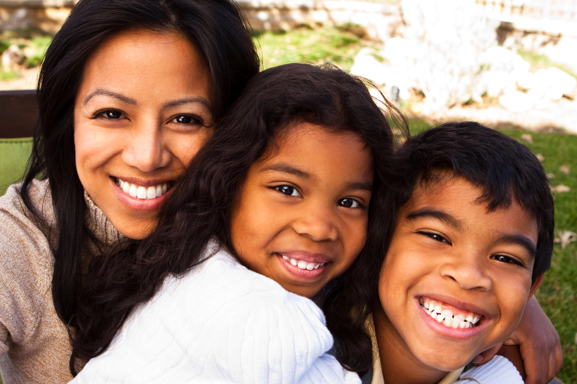 A smiling woman embraces two young children, a boy and a girl, in an outdoor setting. They are all wearing light sweaters and have dark hair. The background features greenery and soft sunlight, creating a warm and cheerful atmosphere.