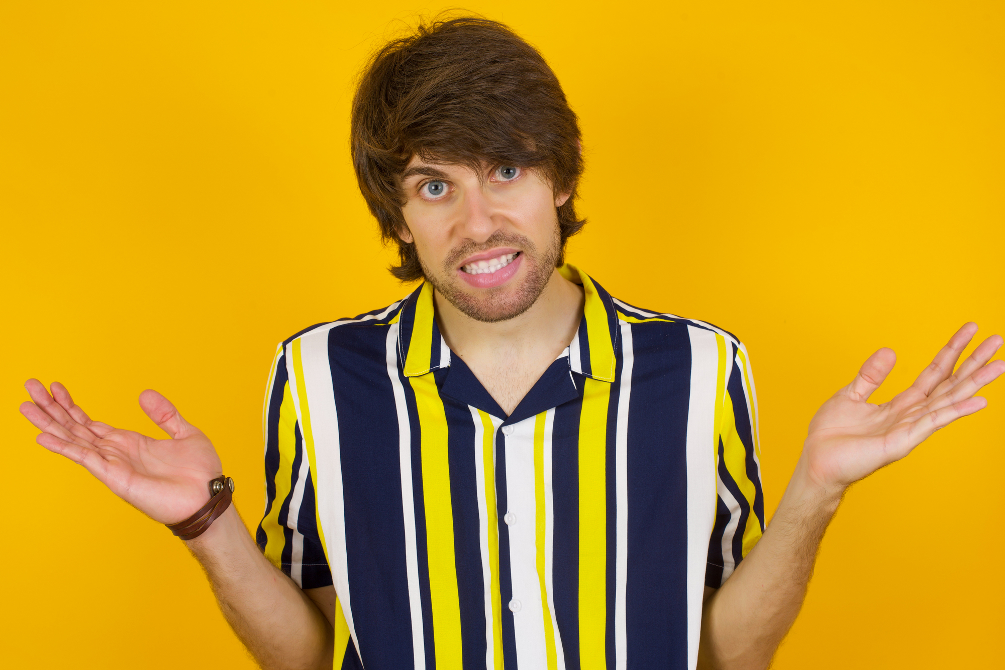 A person with brown hair wearing a yellow, white, and black striped shirt shrugs with their palms up against a bright yellow background.