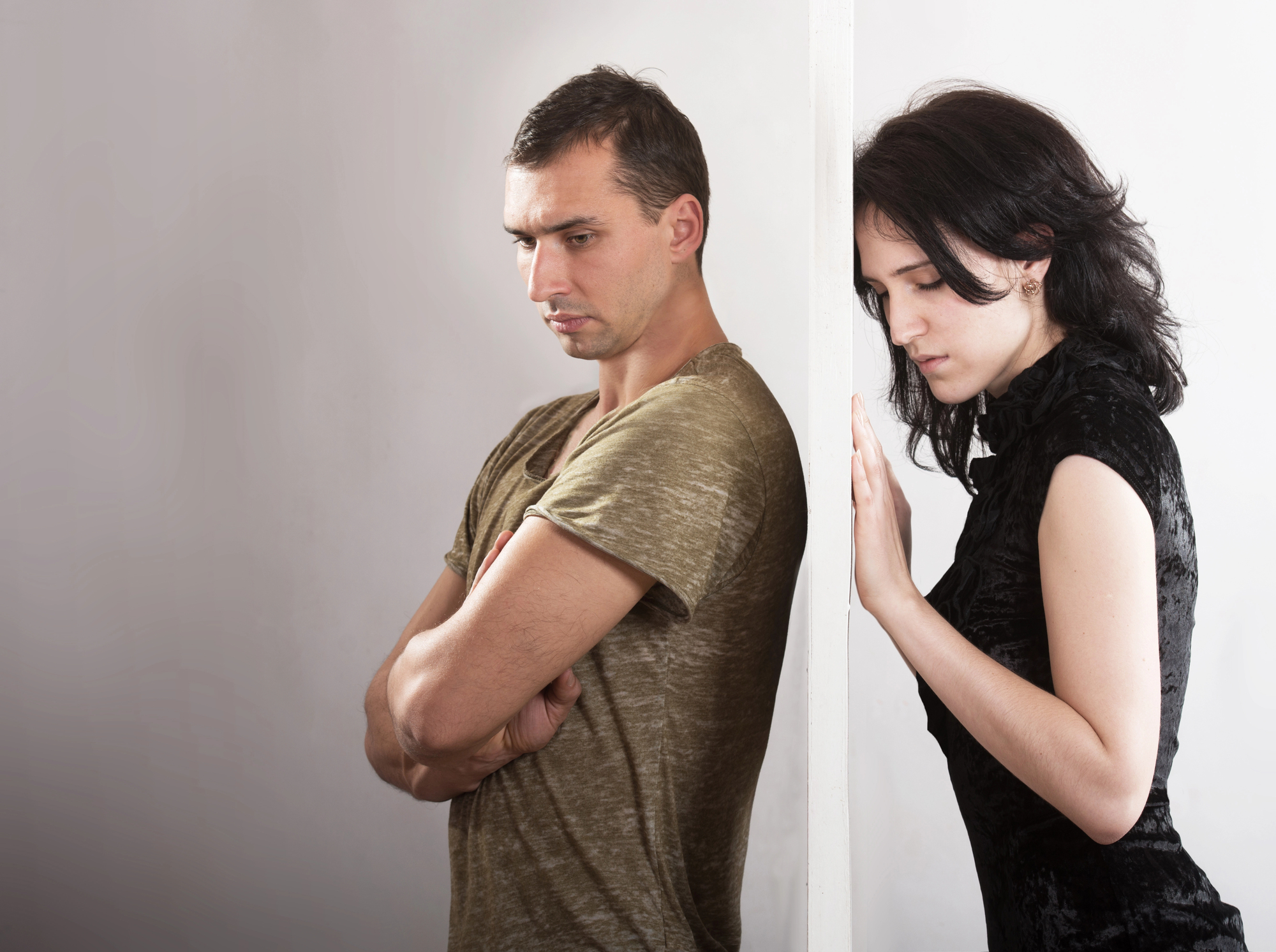 A man and a woman stand back-to-back, separated by a white wall. The man is in a green shirt with arms crossed, looking down. The woman in a black top leans against the wall, appearing thoughtful. The background is neutral.