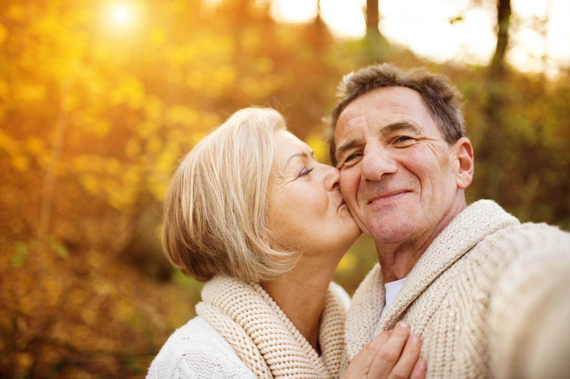 An older couple posing for a selfie in a forest with autumn leaves. The woman is kissing the man on the cheek, both wearing cozy sweaters and smiling. Sunlight filters through the trees, creating a warm, golden atmosphere.