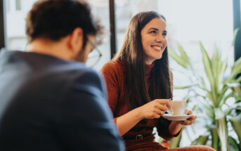 A woman with long dark hair sits holding a cup and saucer, smiling and engaged in conversation. She wears a rust-colored dress. A blurred man in a suit is in the foreground. Green plants and large windows are in the background.