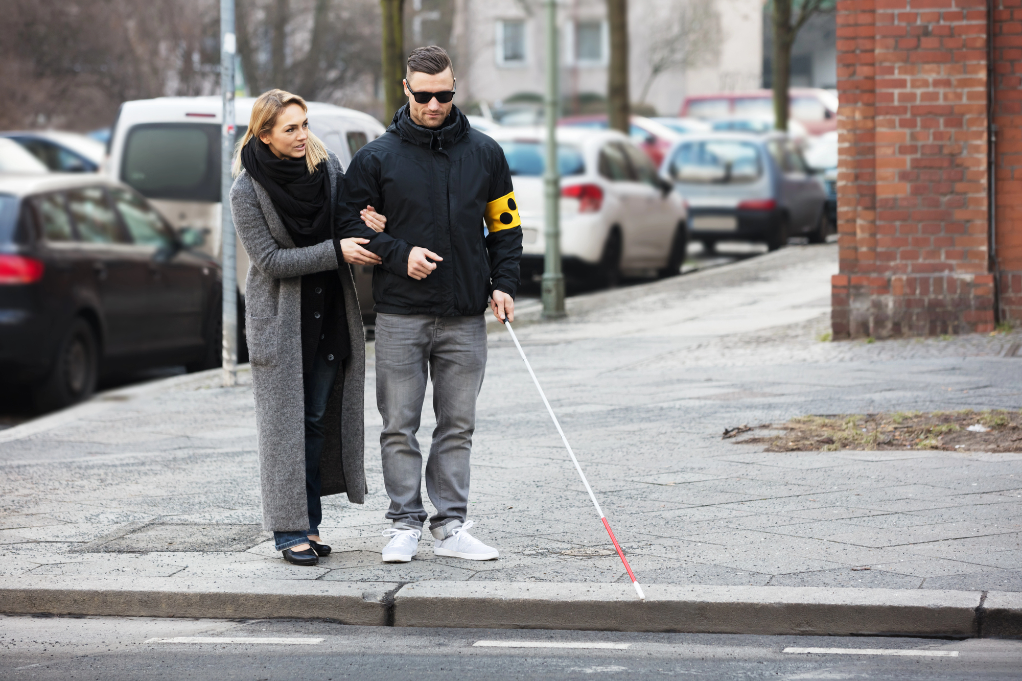 A woman guides a blind man with a white cane as they walk on a city sidewalk. He wears a jacket with a yellow armband and sunglasses, and she is dressed in a long gray coat and scarf. Cars are parked in the background.