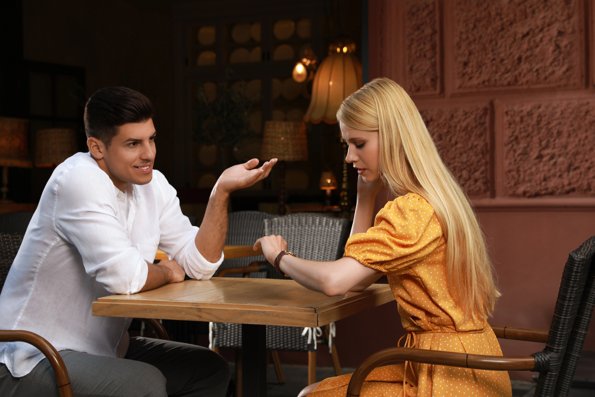 A man in a white shirt sitting at a table gestures while speaking to a woman in a yellow dress who is looking at her watch. They are outdoors at a cafe, surrounded by wicker chairs and warm lighting. The mood seems casual yet serious.