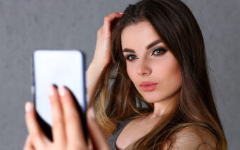 A woman with long brown hair takes a selfie with a smartphone. She has striking makeup, including bold eyeliner and lipstick, and is wearing a sleeveless top. The background is a plain, neutral color.