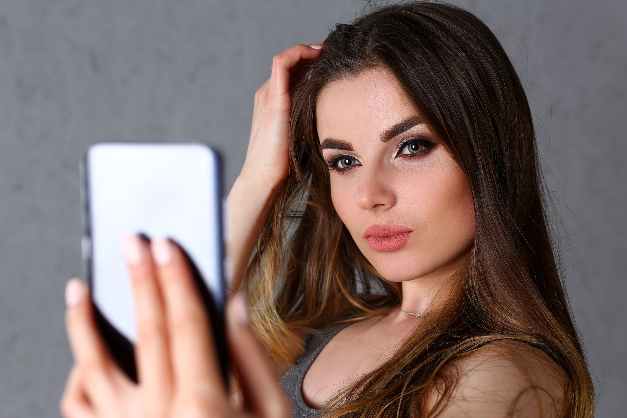 A woman with long brown hair takes a selfie with a smartphone. She has striking makeup, including bold eyeliner and lipstick, and is wearing a sleeveless top. The background is a plain, neutral color.