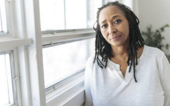 A woman with long, dark braided hair wearing a white blouse stands by a window, looking slightly to the side. Sunlight filters through, casting a soft glow, with greenery visible in the background.