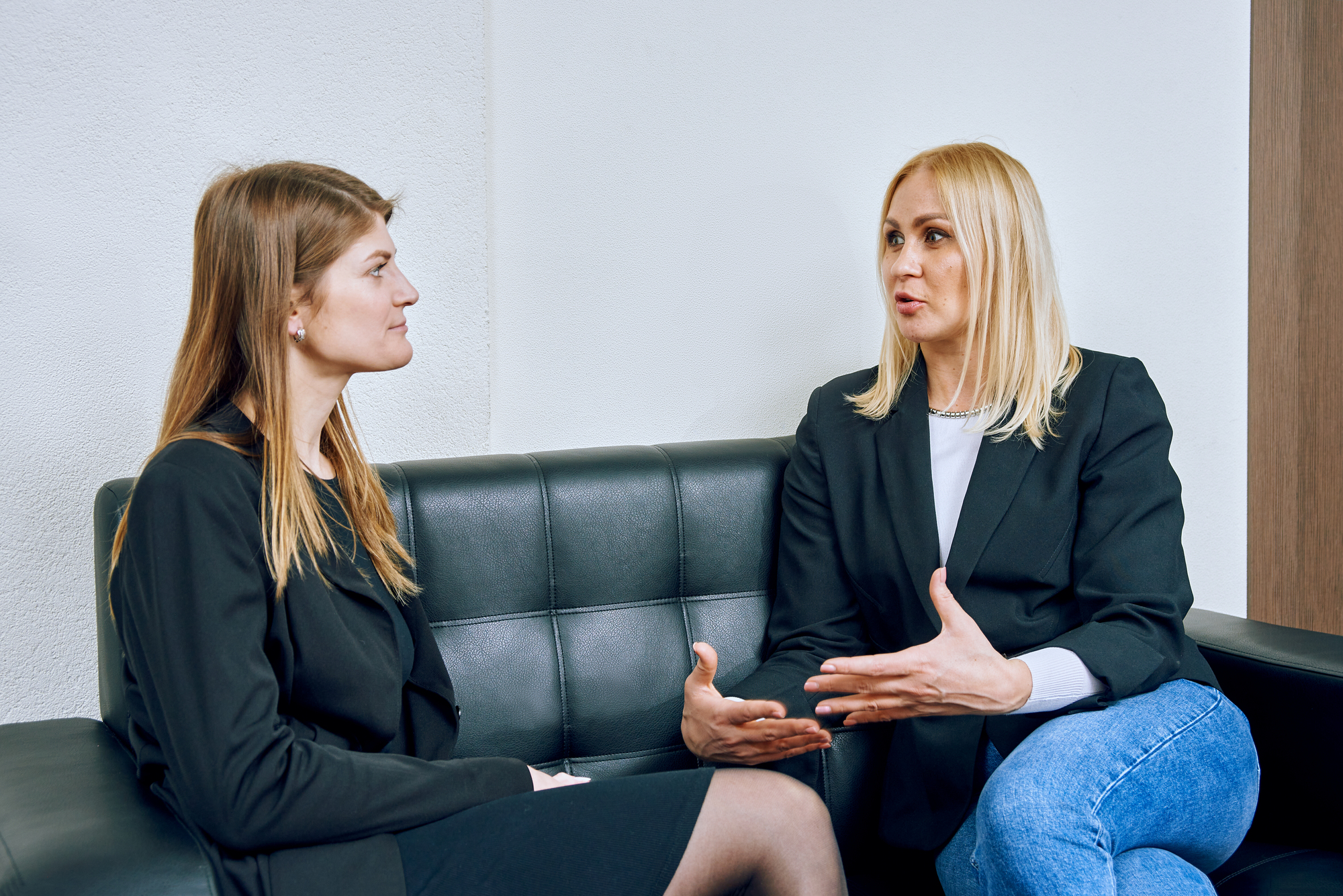 Two women are sitting on a black leather sofa having a conversation. They are both wearing dark blazers, and one is gesturing with her hands. The setting appears to be a professional environment.