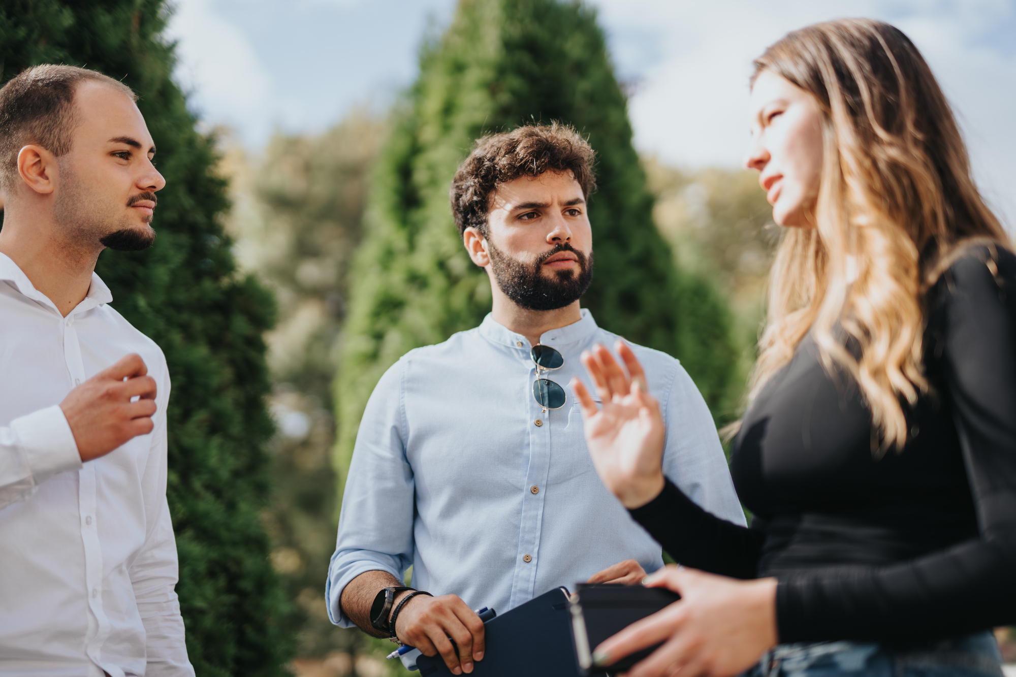 Three individuals are engaged in conversation outdoors. A woman gestures while speaking, and two men listen intently. They stand in front of tall, green trees, enjoying a bright day. One man holds a tablet in his hand.